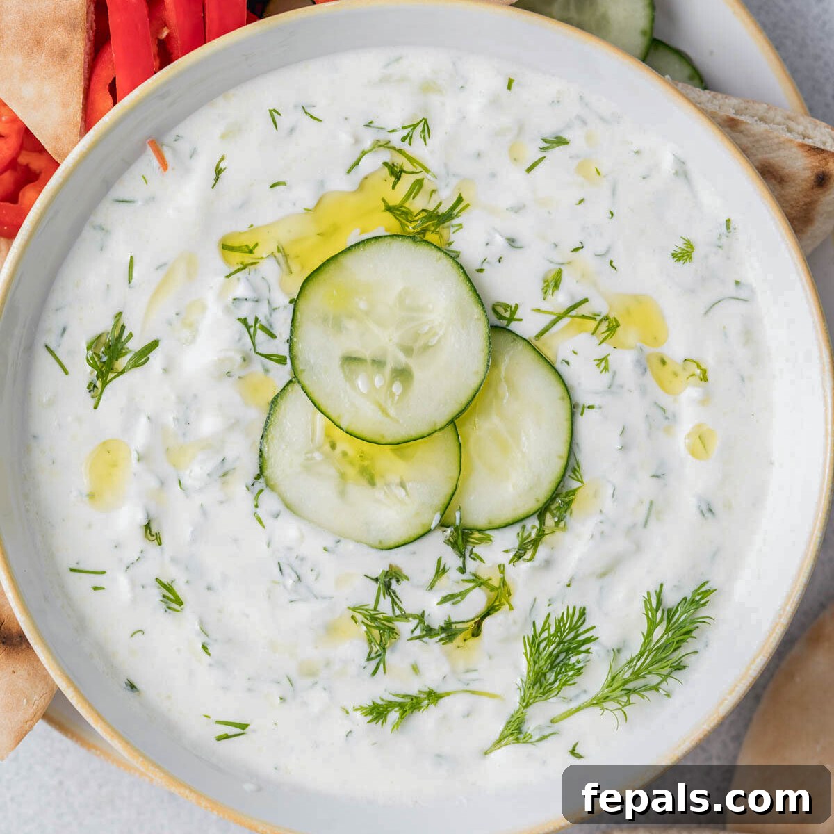 A large bowl of creamy Greek Tzatziki, topped with thinly sliced cucumbers arranged artfully and a generous drizzle of golden olive oil, ready to be served.