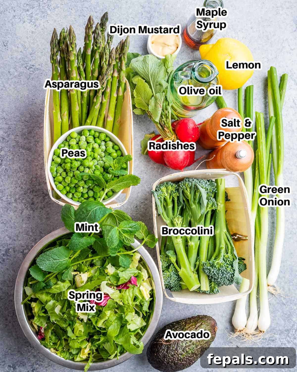 A collection of fresh ingredients laid out on a white surface, ready to make a spring mix salad, including greens, broccoli, asparagus, radishes, peas, avocado, green onions, and fresh mint.