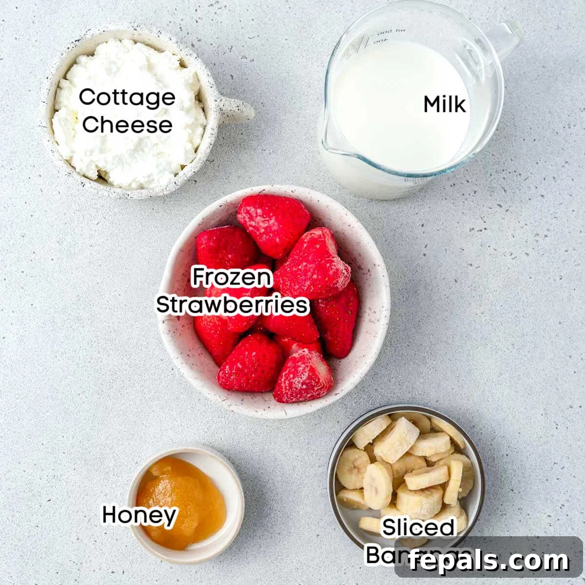 A selection of fresh and frozen ingredients laid out for making a strawberry cottage cheese smoothie: a container of low-fat cottage cheese, a carton of vanilla almond milk, a bag of frozen strawberries, a ripe banana, and a jar of honey.