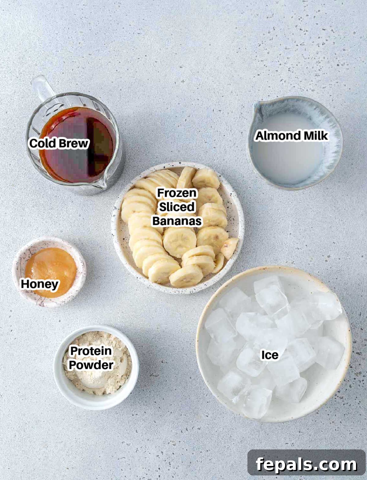 Brewed Protein Fuel 3 A flat lay of all ingredients laid out for a coffee protein shake: frozen banana slices, cold brew coffee, almond milk, vanilla protein powder, ice, and a small jar of honey.
