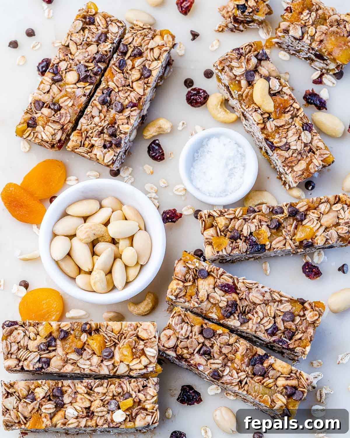 Healthy granola bars spread out on a white surface, surrounded by bowls of dried fruit, and nuts.