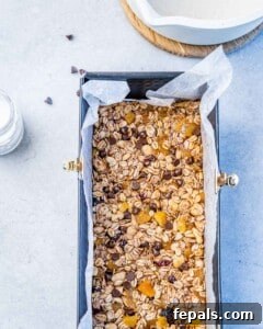 Rectangular loaf pan lined with parchment paper filled with pressed granola mixture with visible oats, nuts, dried fruit, and chocolate chips on top.
