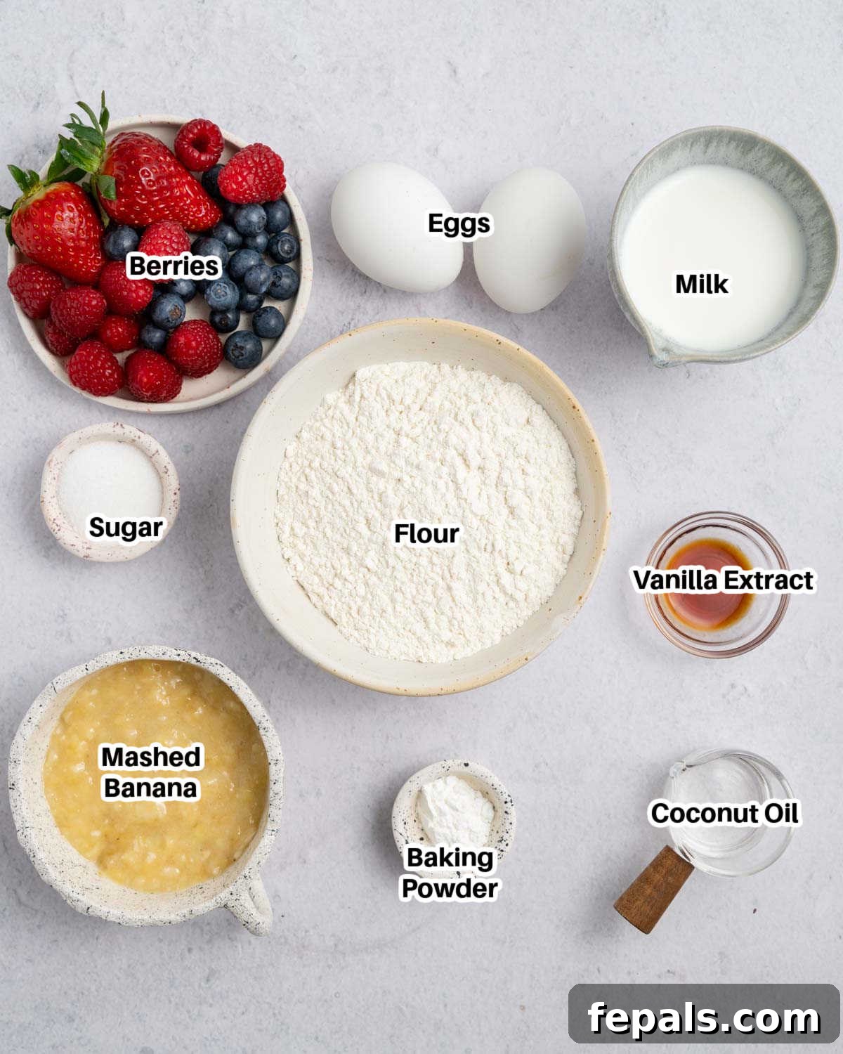 A collection of ingredients laid out on a kitchen counter, including a ripe banana, eggs, flour, milk, and a bowl of fresh berries, ready for making mini pancake muffins.
