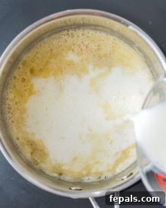 Warm milk being slowly poured into the butter and flour roux in a pan, as part of the Bechamel sauce preparation process.