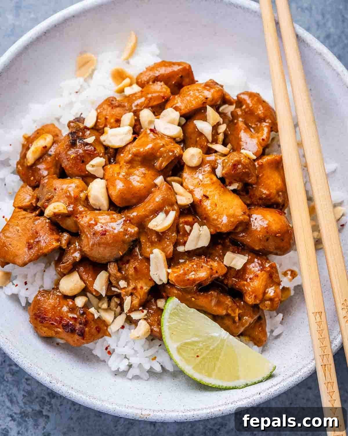 A white bowl and chopsticks with rice, a lime wedge, and peanut butter chicken.