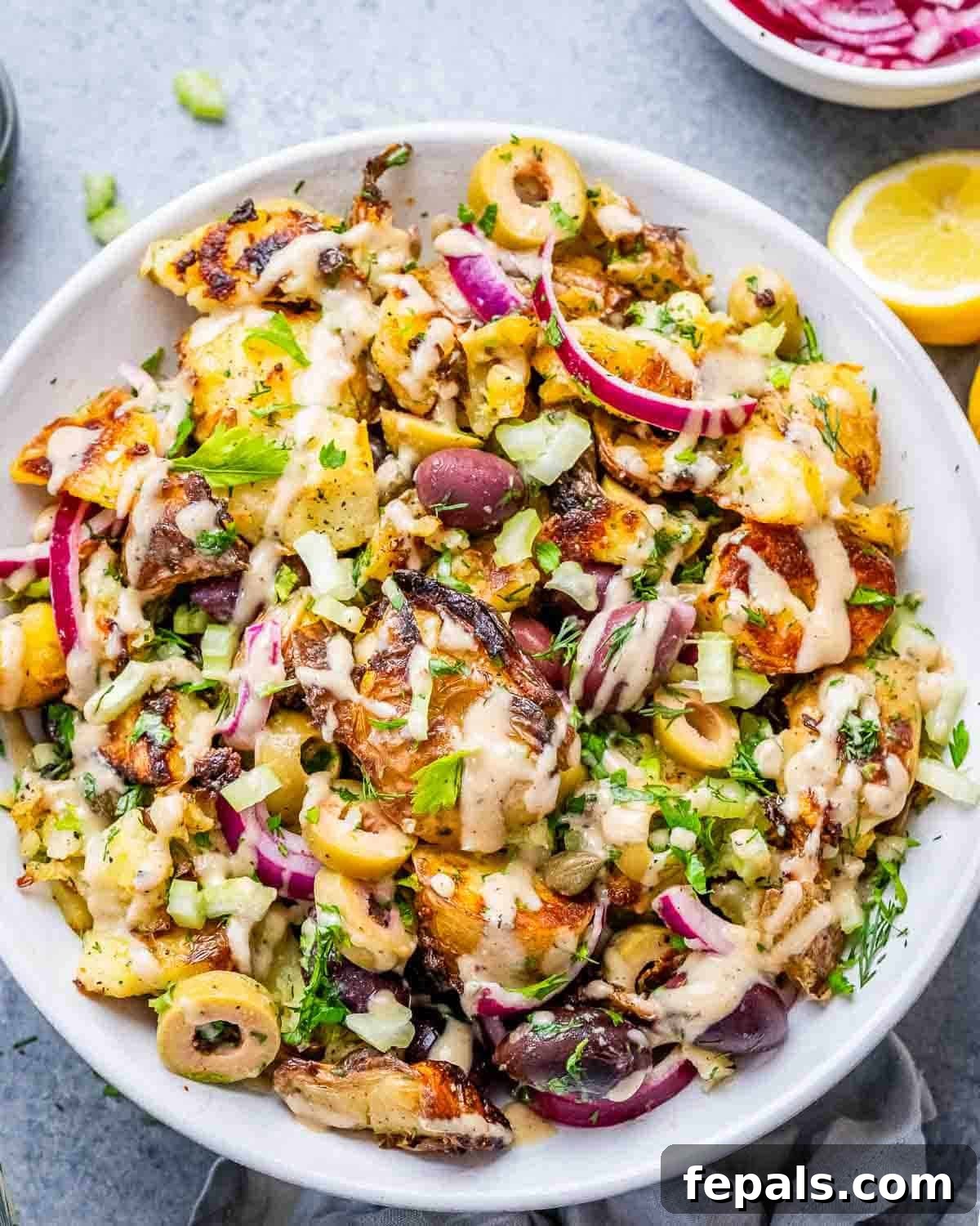 A large white serving bowl filled with crispy smashed potato salad, olives, fresh herbs, and a creamy tahini dressing.