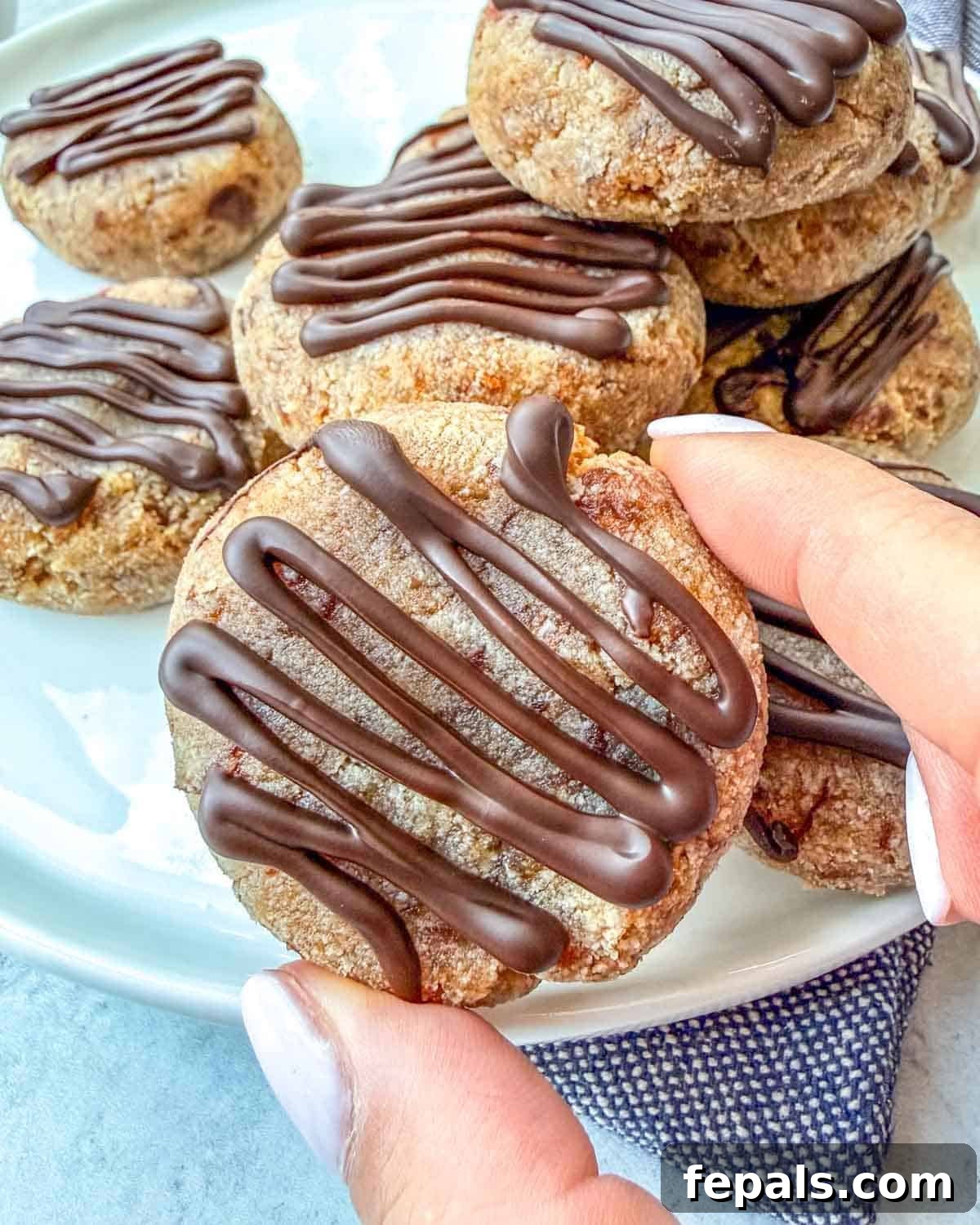 A hand holding a single, delicious almond flour date cookie with a chocolate drizzle.