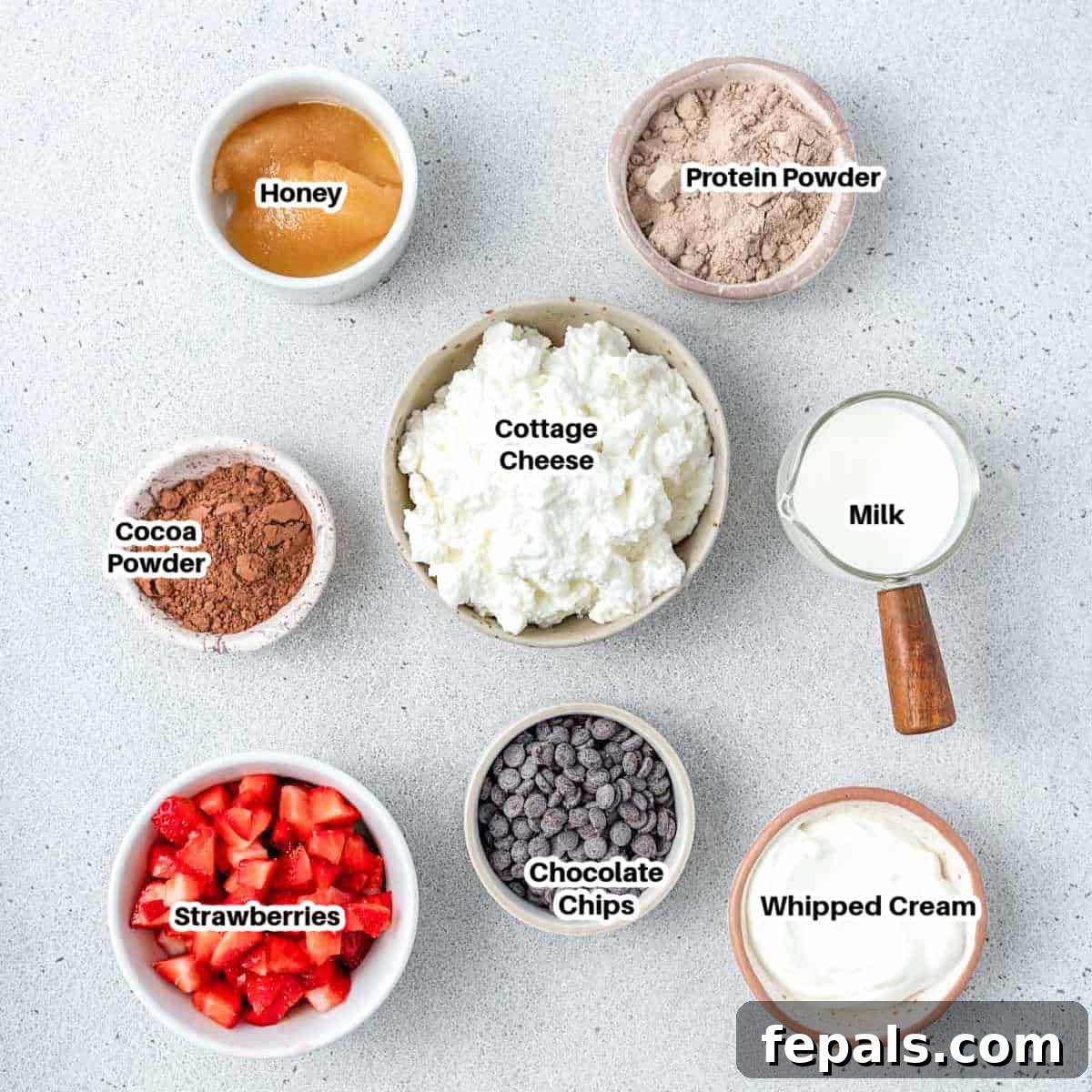 The ingredients for cottage cheese chocolate pudding arranged on a counter: cottage cheese, cocoa powder, protein powder, honey, milk, and chocolate chips.