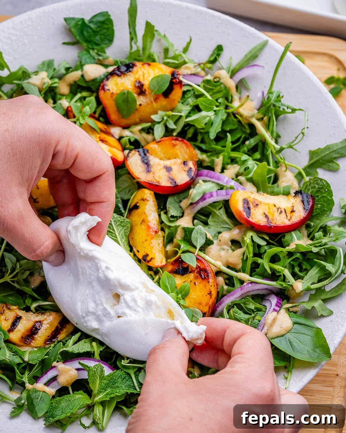Hands tearing burrata over a peach salad.
