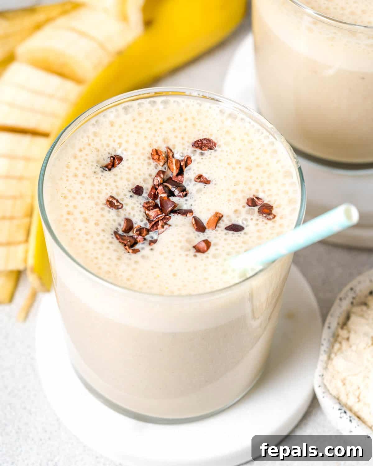 a glass filled with banana protein shake topped with cocoa nibs and a straw in the cup.
