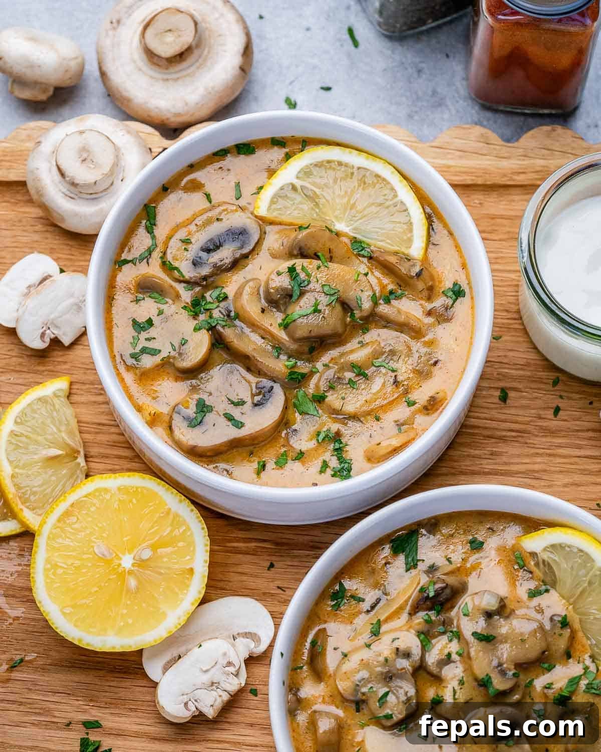 Two white bowls of Hungarian mushroom soup on a wooden board, with a wedge of lemon.