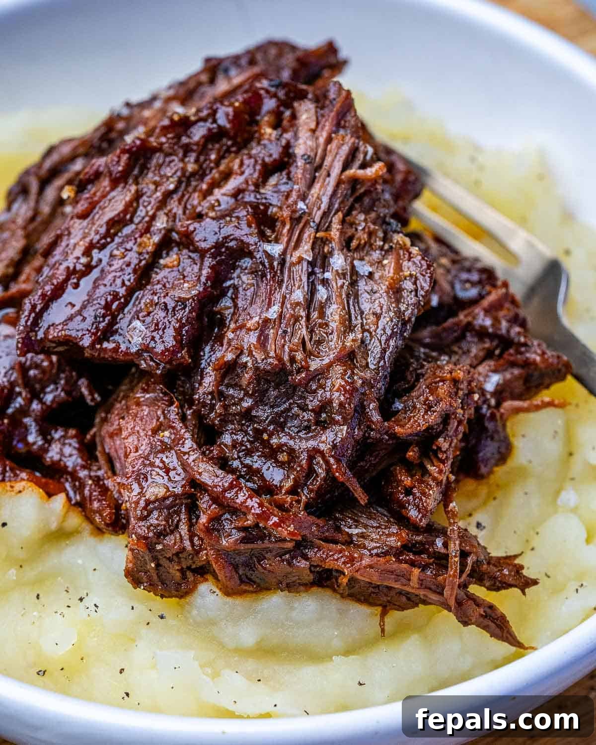 A white bowl of mashed potatoes topped with BBQ slow cooker chuck roast.