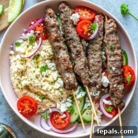 top view of grilled kafta skewers on a round plate with hummus, fresh tomatoes, and crumbled feta cheese