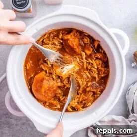 Hand shredding cooked BBQ chicken with two forks inside a white crockpot.